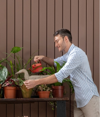 homem regando plantas parede madeira marrom escuro Homem regando plantas em frente a uma parede de madeira de tom marrom escuro.