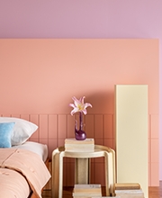 lirio quarto violetas Quarto tem cama arrumada, uma mesa de cabeceira apoiando um vaso e sua flor e, ao fundo paredes pintadas em tons calmos de violetas e rosas.